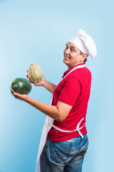 Handsome indian male chef holding water melon and musk melon, asian male chef holding fruits, isolated over blue background