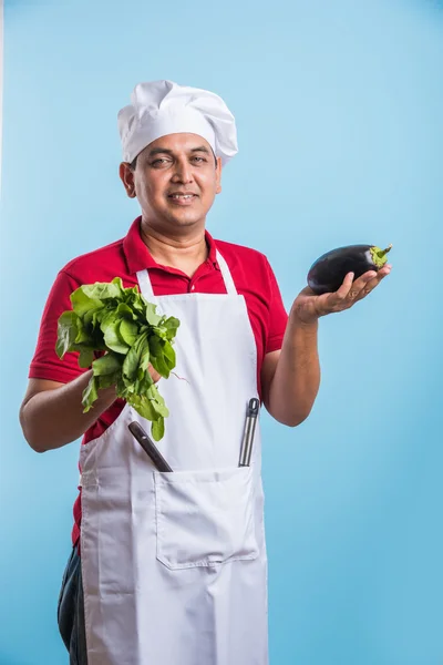 Indian male chef holding fresh Cauliflower and spinach leaves, standing isolated over blue background, asian male chef holding fresh vegetables
