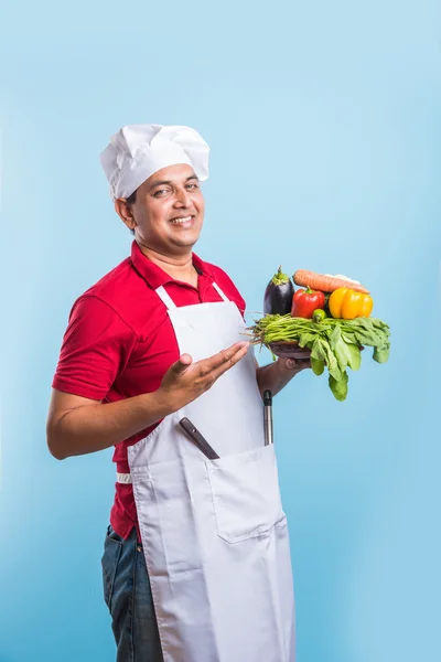 Indian male chef holding fresh vegetables in hand, asian male cook chef standing with vegetables, isolated over blue background