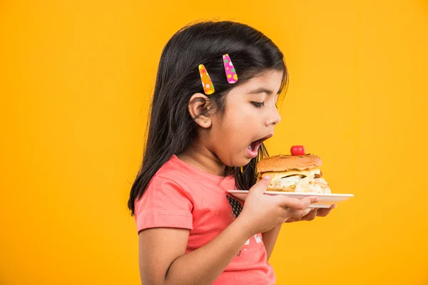 Cute indian girl eating burger, small asian girl and burger, isolated over yellow background