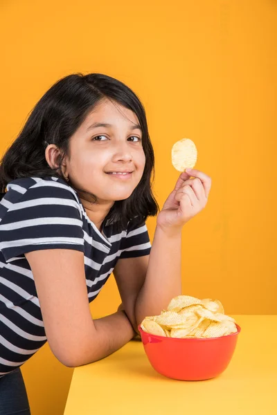 Cute little indian girl eating chips or potato wafers, asian girl eating potato chips, small girl eating chips in red bowl, over yellow background