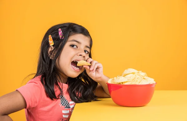 Cute little indian girl eating chips or potato wafers, asian girl eating potato chips, small girl eating chips in red bowl, over yellow background