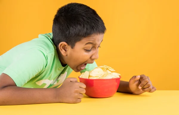 Cute little indian boy eating chips or potato wafers, asian boy eating potato chips, small boy eating chips in red bowl, over yellow background