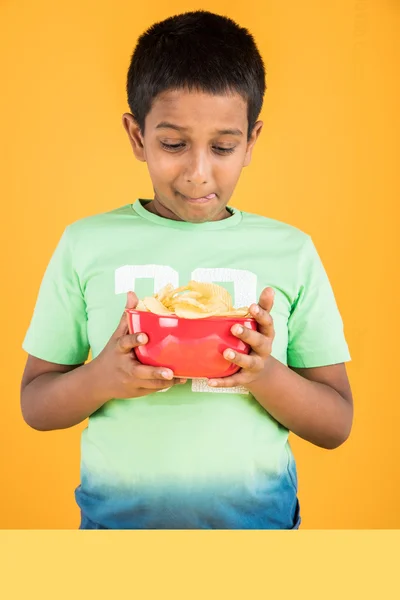 Cute little indian boy eating chips or potato wafers, asian boy eating potato chips, small boy eating chips in red bowl, over yellow background