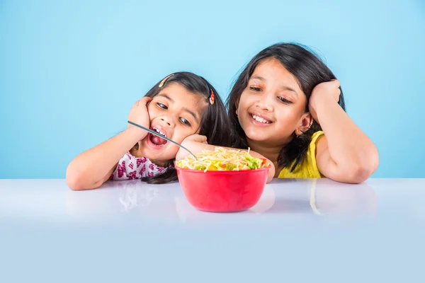 Happy Asian children eating delicious noodle, two cheerful little indian girls eating noodles in red bowl over blue background, two indian little sisters or friends with noodles