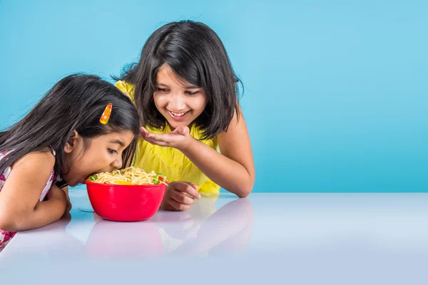 Happy Asian children eating delicious noodle, two cheerful little indian girls eating noodles in red bowl over blue background, two indian little sisters or friends with noodles