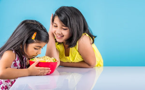 Happy Asian children eating delicious noodle, two cheerful little indian girls eating noodles in red bowl over blue background, two indian little sisters or friends with noodles