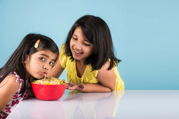 Happy Asian children eating delicious noodle, two cheerful little indian girls eating noodles in red bowl over blue background, two indian little sisters or friends with noodles