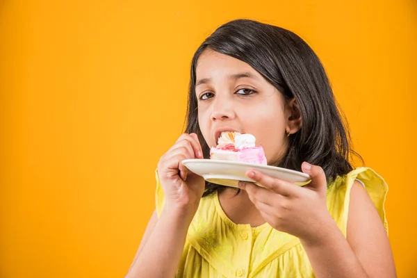 Portrait of Indian kid eating cake or pastry, cute little girl eating cake, girl eating strawberry cake over yellow background