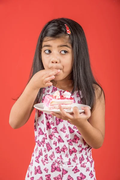 Portrait of Indian kid eating cake or pastry, cute little girl eating cake, girl eating strawberry cake over red background