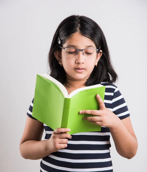 Indian small girl with eye glasses standing and reading a book, asian little girl holding a book and reading over white background, cute indian girl wearing spectacles and reading book while standing