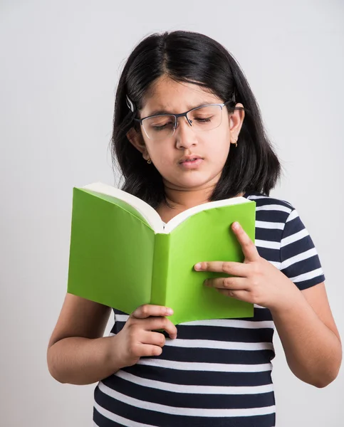 Indian small girl with eye glasses standing and reading a book, asian little girl holding a book and reading over white background, cute indian girl wearing spectacles and reading book while standing