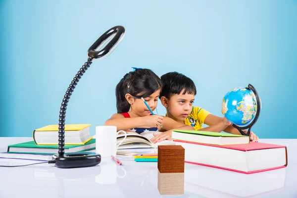 Indian kids studying geography and mathematics, small asian siblings studying, indian girl and boy doing homework on study table with globe, milk mug and pile of books, indian kids writing