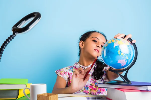 Indian girl student looking at educational globe and sitting at table with books, table lamp and milk mug, asian girl child studying geography, curious asian girl studying geography with globe