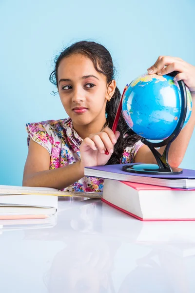 Indian girl student looking at educational globe and sitting at table with books, table lamp and milk mug, asian girl child studying geography, curious asian girl studying geography with globe