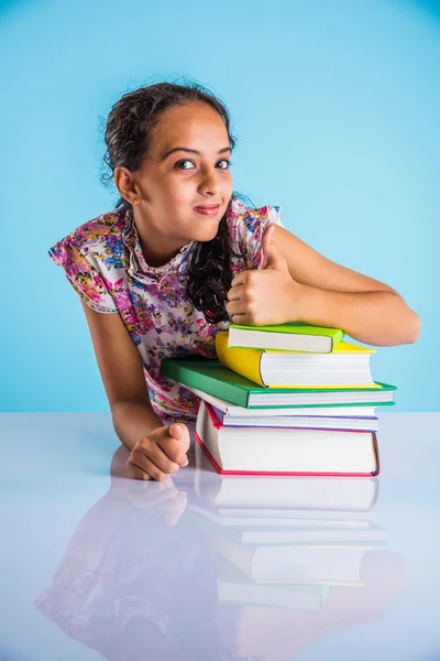 Indian studious girl showing gold medal or success sign, leaning over table with pile of books, asian small girl and success concept, 1st in studies concept, indian schoolgirl and merit concept