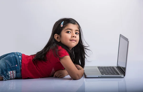 Cheerful indian little girl using laptop, asian small girl playing on laptop, isolated over white background, cute indian baby girl lying on floor playing on laptop