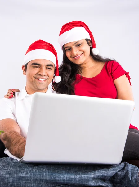 Portrait of beautiful indian couple with laptop in christmas time. Christmas tree in background. indian family & christmas time, indians with red santa hat, wife pointing to laptop, shopping, surfing
