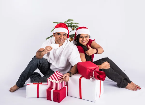 Young indian couple sitting with with christmas gifts showing card, asian couple and christmas gift, with santa hat, white background, india and christmas celebration, christmas gift and indian couple