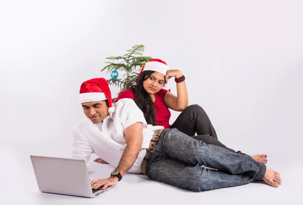 Young indian couple with red santa hat, sitting close on white background with christmas tree, man busy on laptop and wife upset, busy man and sad wife on christmas time