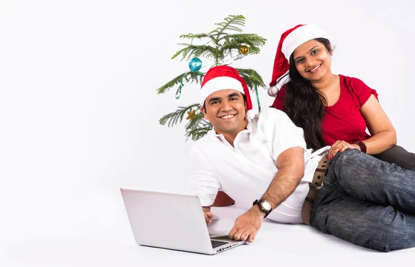 Portrait of beautiful indian couple with laptop in christmas time. Christmas tree in background. indian family & christmas time, indians with red santa hat, wife pointing to laptop, shopping, surfing