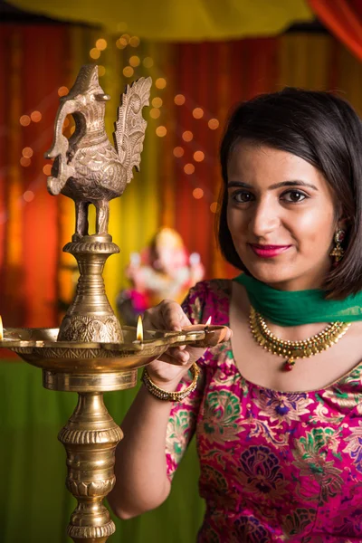 Indian young and beautiful girl in traditional wear lighting oil lamp or samai with diya and celebrating ganesh festival or Diwali or deepavali. Indian girl hands holding oil lamp indoors.