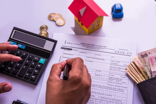 Indian man or accountant person filing Indian income tax returns form or ITR document showing indian currency, house model, toy car and calculator over white table top, selective focus