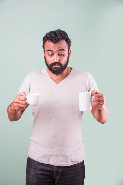 Stylish Indian man with beard with two cups of coffee in two hand looking at camera.