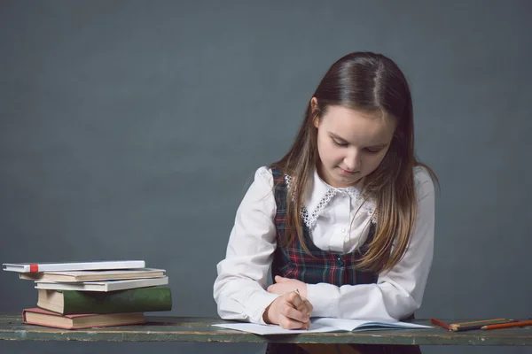 Girl in a school uniform sitting at a table with books, gray background, looks like a chalk board