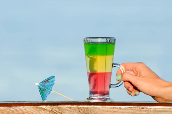 Female hand holding a glass with colorful cocktail on the background of the sea, umbrella for cocktails