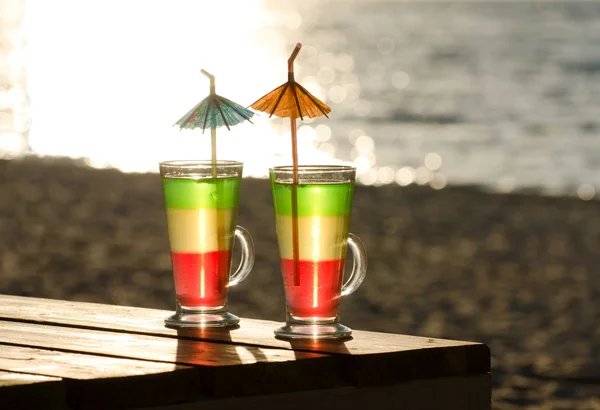 Glasses with multi-colored cocktails on a wooden table in the sun, the beach and the sea in the background