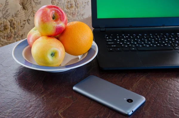 Apples and oranges on a plate next to a laptop, smart phone on the table. Healthy break