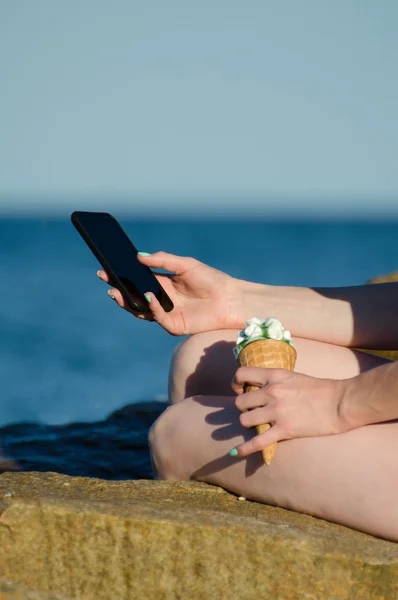A girl sits on a rock near the sea with a smartphone and ice cream in the hands of