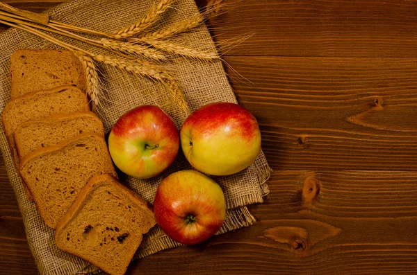 Slices of rye bread, three apples, ears of wheat on sacking, wooden table, top view