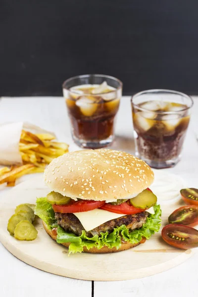 Burger with meat, lettuce, tomatoes, cheese, pickles, onions, french fries and sparkling water on a white wooden background