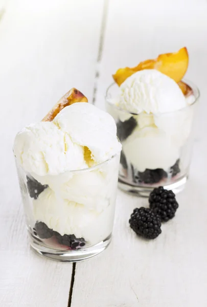 Ice cream in glass with a peach and blackberry on a white wooden background