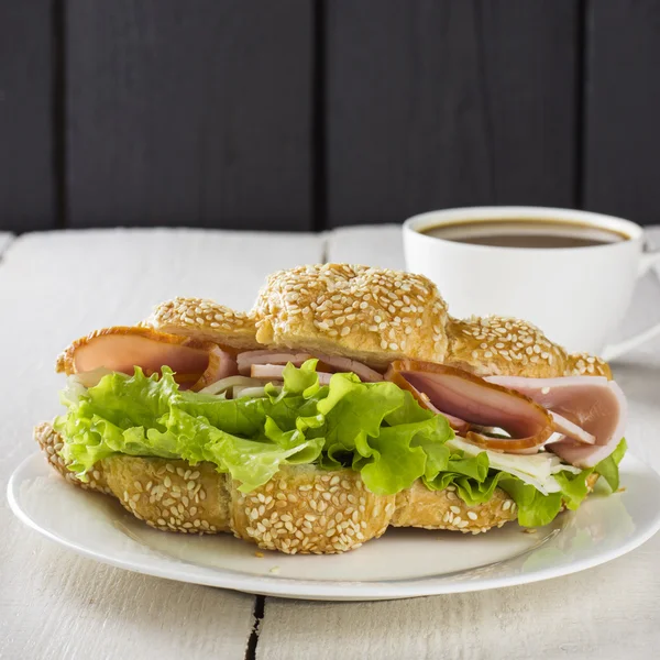 Croissant sandwich with lettuce, bacon, cheese on white plate and cup of coffee on a wooden backgroundCroissant sandwich with lettuce, bacon, cheese on white plate and cup of coffee on a wooden background