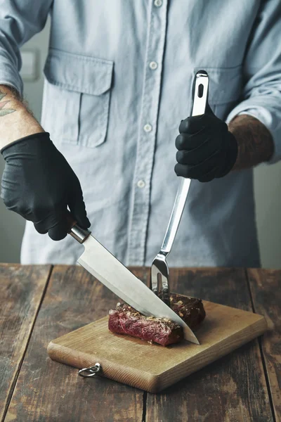 Tattooed butcher hands in black gloves slice grilled meat