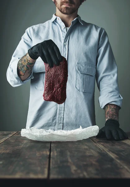 Tattooed butcher holds piece of meat above wooden table