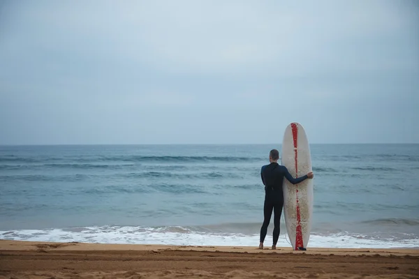 Old surfer with surf board stays alone on beach