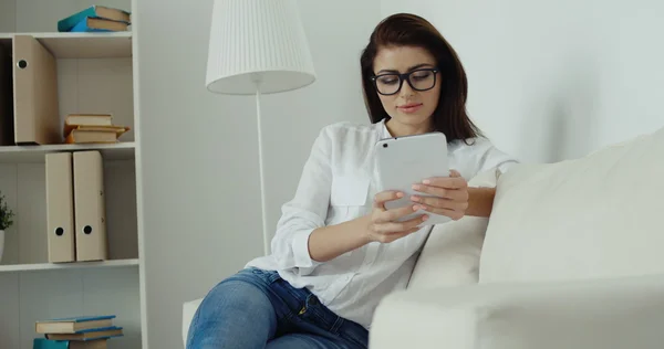 Young brunette businesswoman in white shirt, blue jeans and glasses with computer tablet siting on the white sofa.