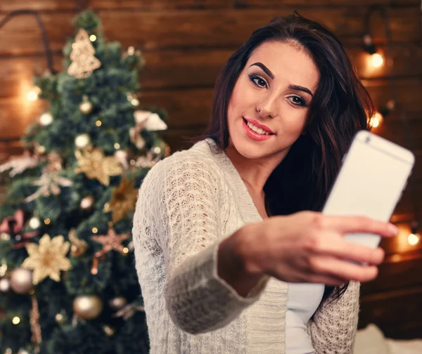 People, holidays and technology concept - beautiful brunette woman in white  sweater taking selfie picture by smartphone over christmas tree and living room background