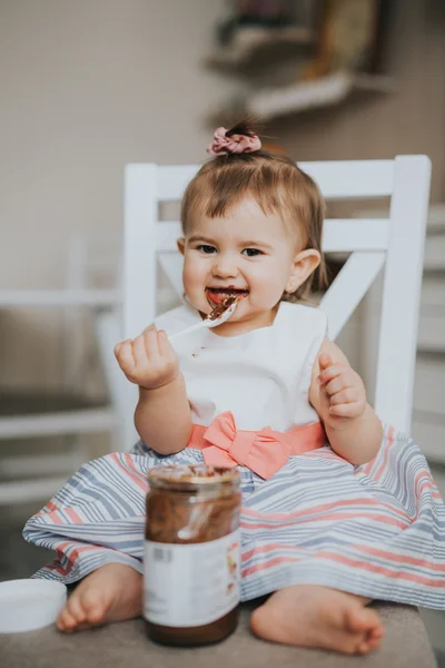 Cute little baby girl on the table in the kitchen eats with the spoon chocolate cream and makes funny happy faces