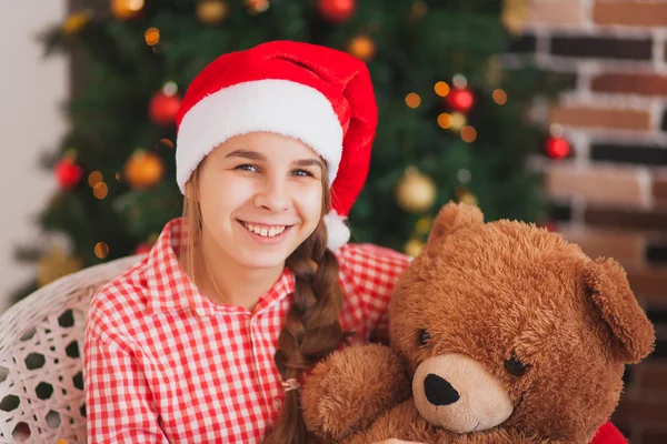 Holidays, presents, christmas, childhood and people concept - smiling girl in santa helper hat with teddy bear over lights background