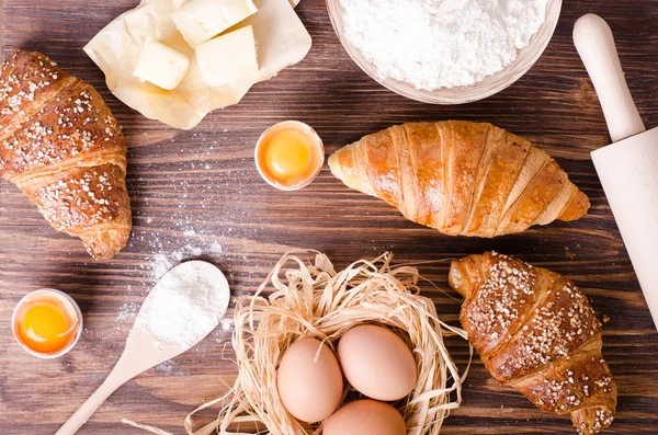 Ingredients for baking croissants - paper, flour, wooden spoon, rolling pin, eggs, egg yolks, butter served on a rustic wooden tray table.