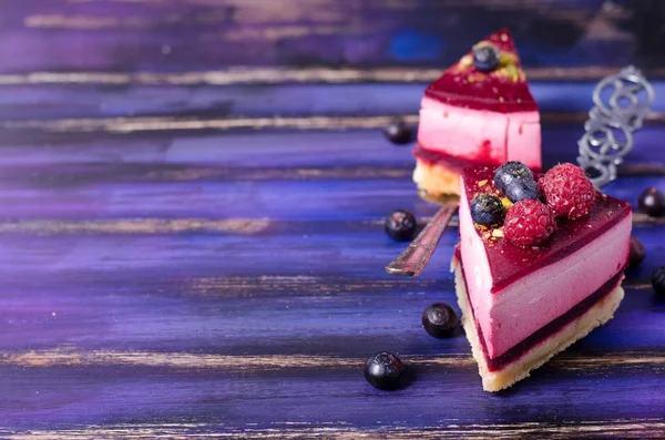 Piece of delicious raspberry cake with fresh raspberries, blueberry, currants and pistachios on shovel, bright purple, violet, blue background. Free space for your text.