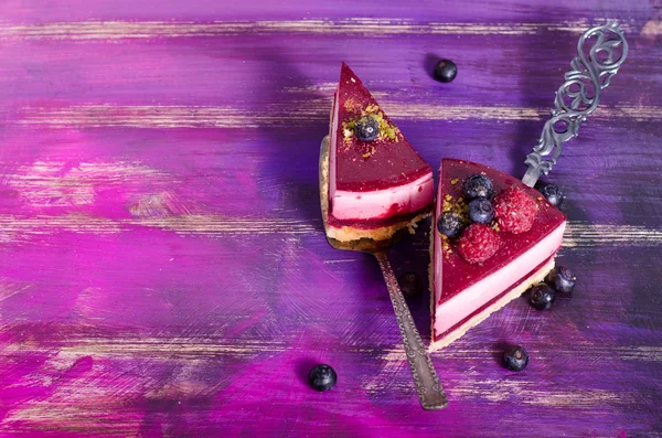 Piece of delicious raspberry cake with fresh raspberries, blueberry, currants and pistachios on shovel, bright purple, violet background. Free space for your text.