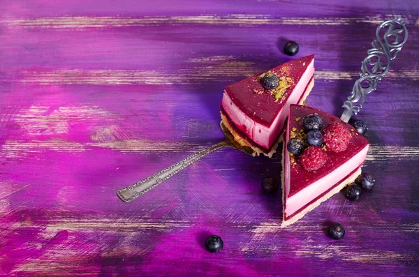 Piece of delicious raspberry cake with fresh raspberries, blueberry, currants and pistachios on shovel, bright purple, violet background. Free space for your text.