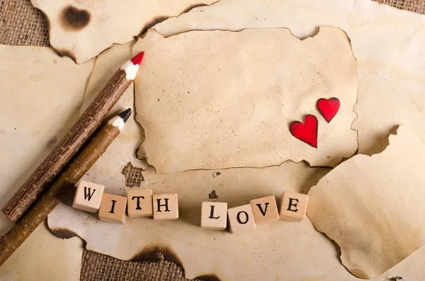 Old vintage sheet of paper, two red hearts, wooden pencils and words With love on cubes on burlap, sackcloth background. Retro design effects.