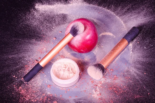 Face powder, two brushes, and red apple on black